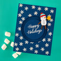 A square navy blue handmade holiday card with a circular cutout in the center and the words 'Happy Holidays' in white script. The card is decorated with white snowflakes and features a paper snowman wearing a red and green hat and brown arms, holding a yellow star.