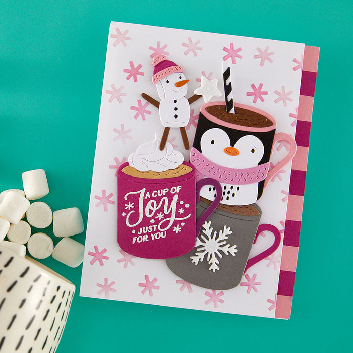 A handmade holiday greeting card featuring cartoon penguins and a snowman with hot cocoa cups, styled on a teal background with marshmallows and a ceramic mug.